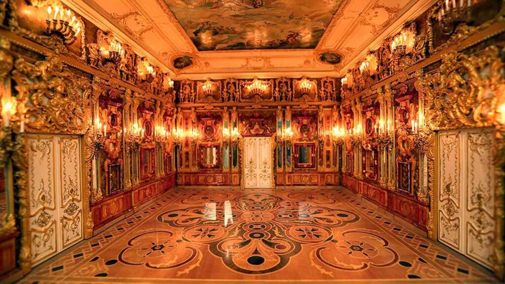 The Amber Room - An ornate, opulent room with elaborate golden wall decorations, intricate woodwork, and glowing chandeliers. The ceiling features detailed artwork, and the polished floor has a geometric pattern.