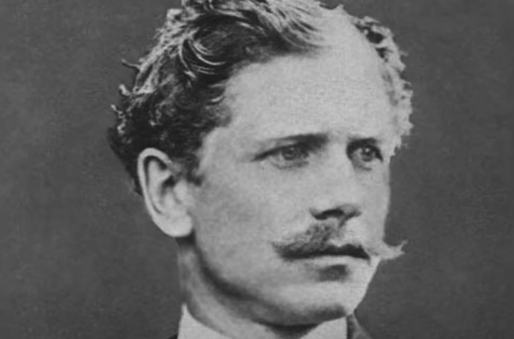 Black and white portrait of a man from the late 19th to early 20th century. He has wavy hair and a mustache, wearing a suit. He gazes to the side with a serious expression.