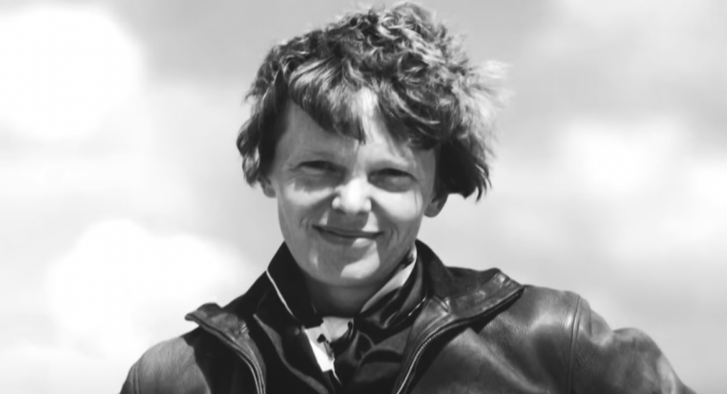 Black and white photo of a smiling person with short, wavy hair, wearing a leather jacket and scarf. The background is lightly blurred, featuring a cloudy sky.