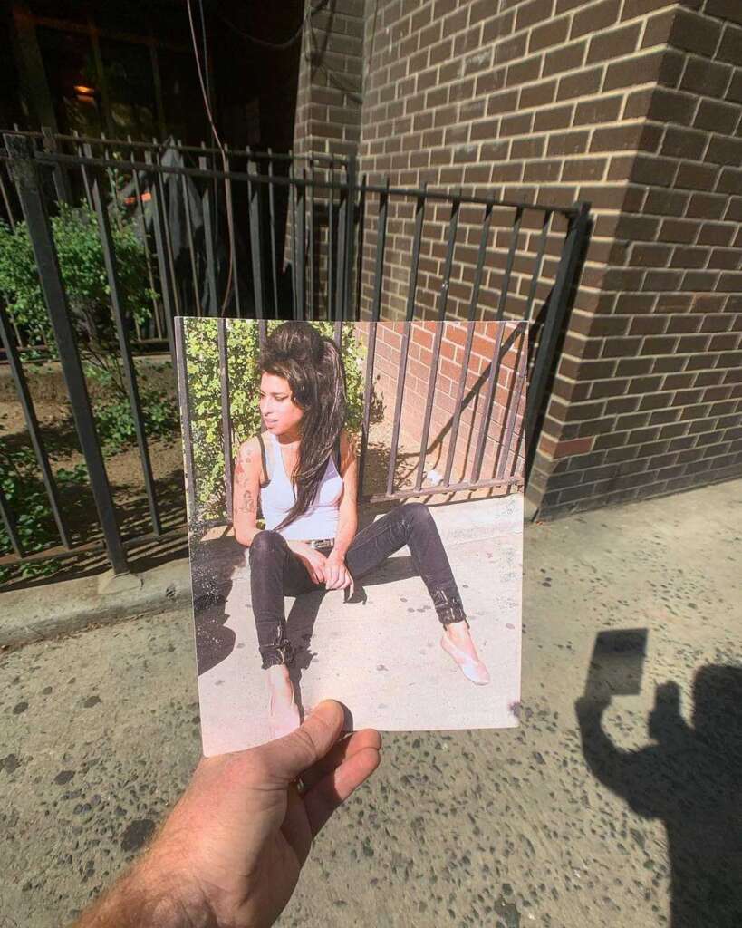 A person holds a photo of a woman sitting on the ground near a brick and iron fence. The woman has dark hair, a white tank top, and black jeans. The shadow of the person holding the photo is visible on the sidewalk.