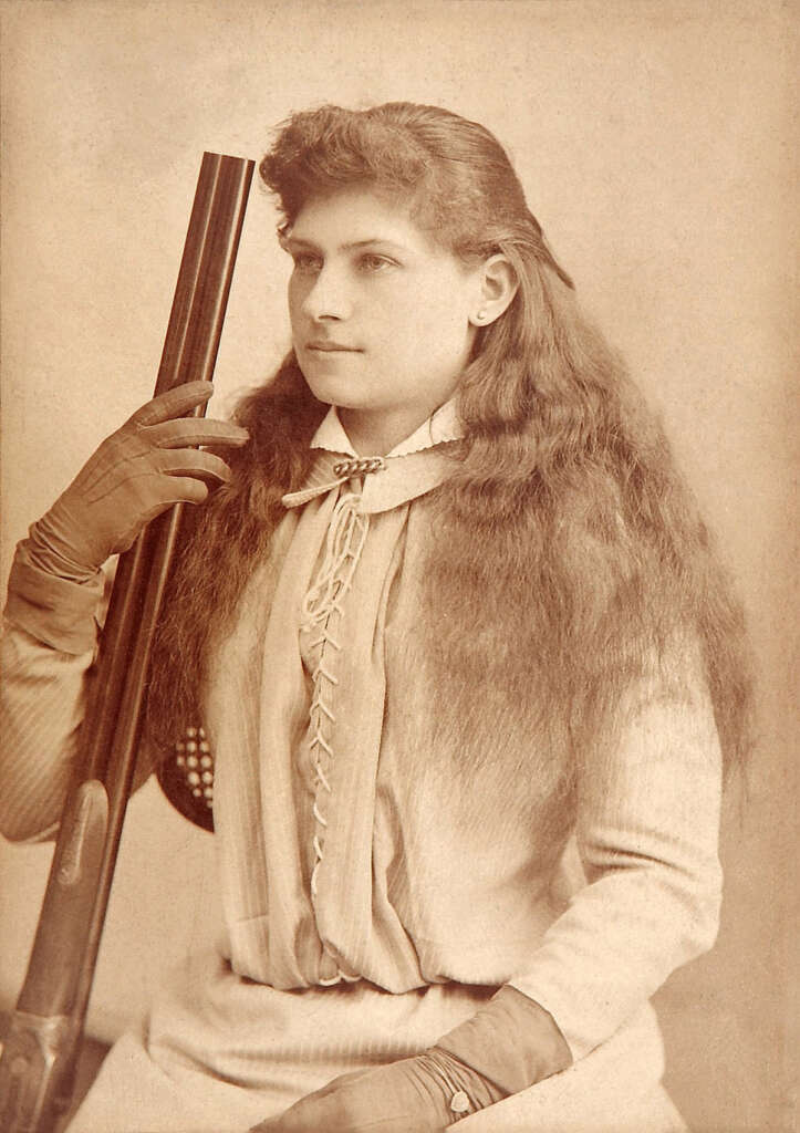 Sepia-toned portrait of a woman with long, wavy hair, dressed in a vintage blouse with a laced front and gloves, holding a rifle. She sits in a dignified pose against a plain backdrop.