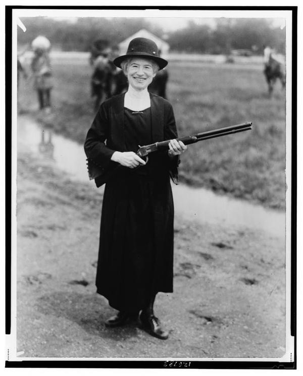 A woman in a long dress and hat stands outdoors holding a shotgun. She is smiling, and the background shows a grassy area with another person slightly blurred in the distance.