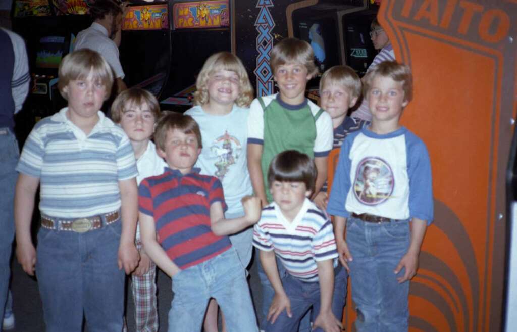 A group of young children stands in front of arcade machines. They are smiling and making various poses. The background features classic arcade games with bright designs. The children are dressed in casual 1980s clothing.