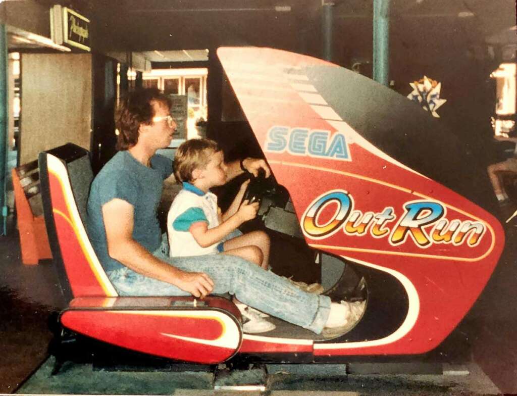 A man and a child sit in a red SEGA OutRun arcade racing game cabinet. The man is in the driver's seat while the child is on his lap, holding the steering wheel. The arcade setting is dimly lit.