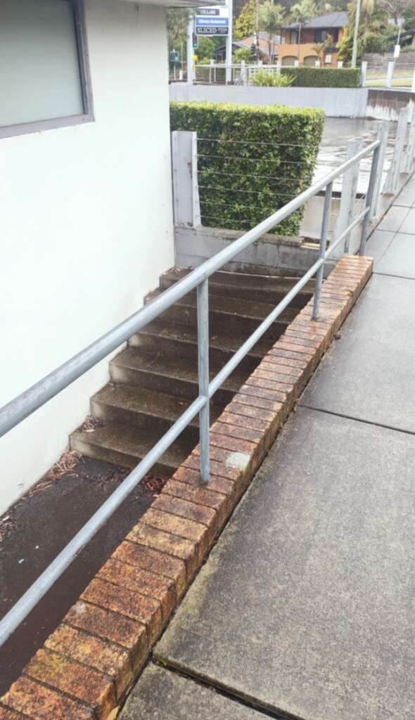 Brick-bordered ramp with metal railing leading up to a building, adjacent to shrubbery and concrete pavement, on a rainy day.