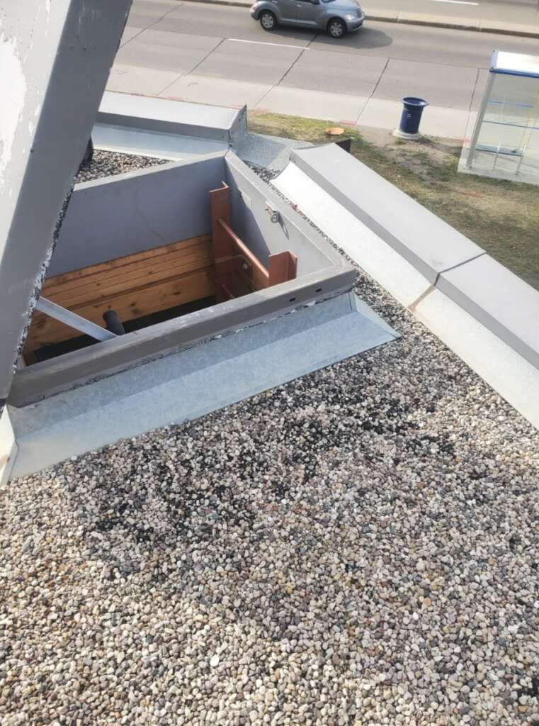 View of a gravel rooftop with an open hatch and some wooden beams inside. A road with a moving car, a bus stop, and a blue trash bin are visible in the background.