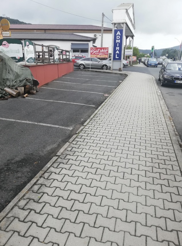 A paved sidewalk runs parallel to an empty parking lot with a few parked cars. A large sign reading "ADMIRAL" is visible near the building in the background. The sky is overcast, and there are some covered items on the left.