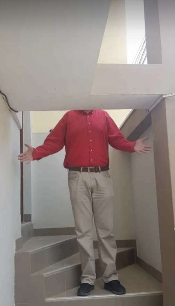 A person standing on stairs with their head obscured by a low ceiling. They are wearing a red shirt and beige pants, and have their arms outstretched.