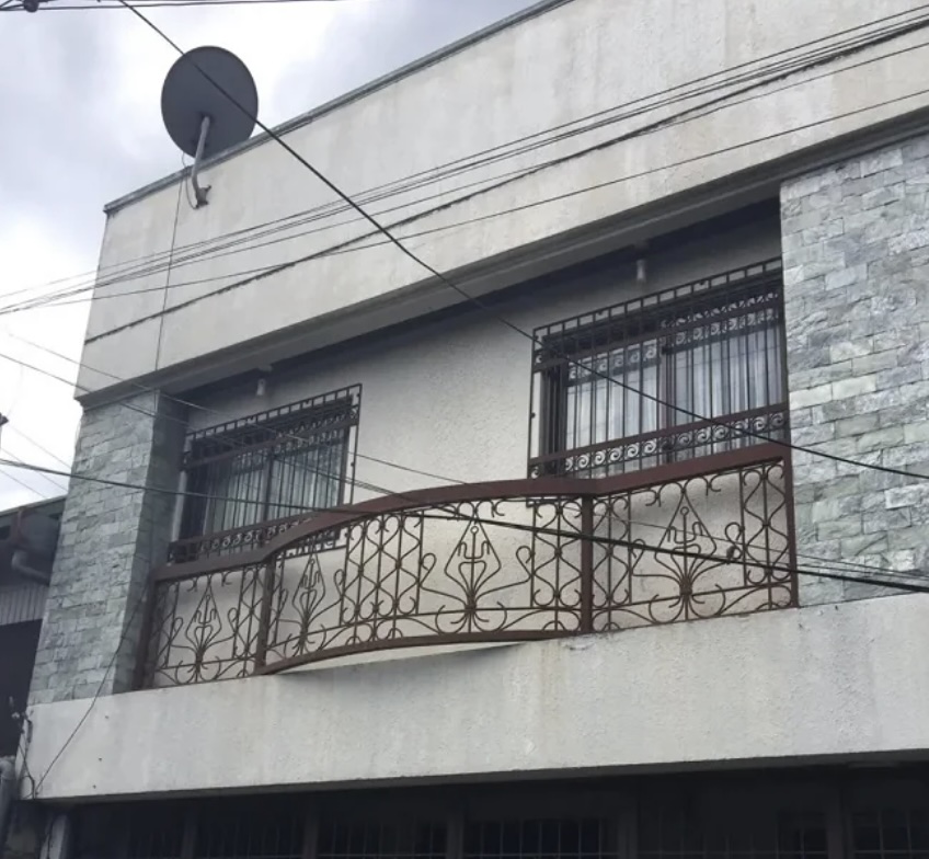 A two-story building facade with two barred windows and a decorative metal balcony. The exterior has a stone-like finish on the right. Overhead wires and a satellite dish are visible against a cloudy sky.