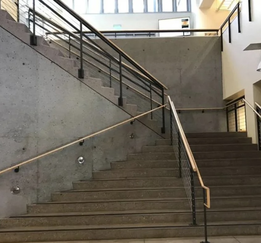 A large, indoor staircase made of concrete with metal railings leads upward, splitting into two directions. The area is well-lit by large windows on the upper floor and ceiling lights.