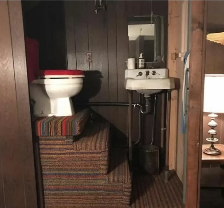 A small bathroom with a toilet elevated on carpeted steps next to a sink. There is a mirror above the sink, with a strip of wall dividing the two areas. A lamp is visible on the right side of the image.