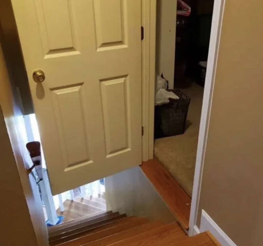 A door opens directly onto a wooden staircase without a landing, leading down to a lower level. A basket with laundry is visible in the adjoining carpeted room.