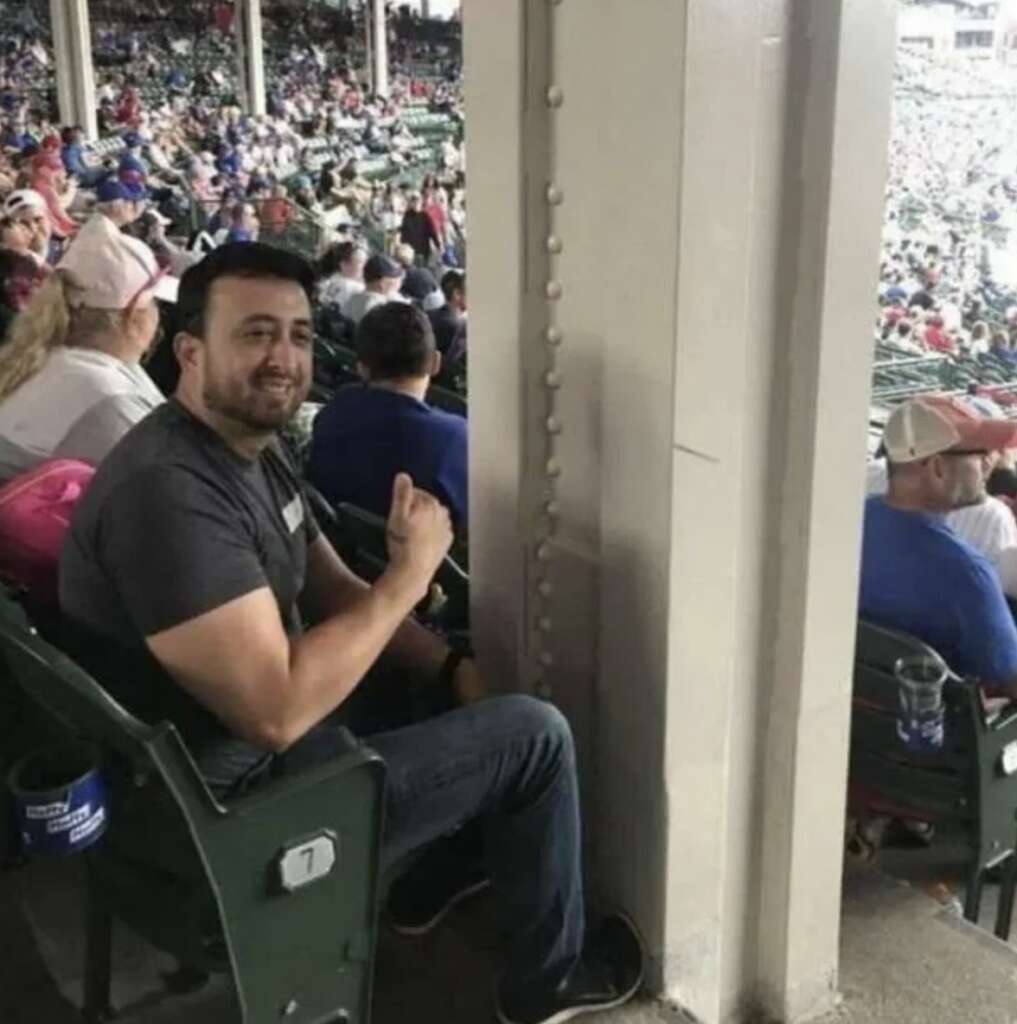 A man sits in a stadium seat, giving a thumbs-up. His view is obstructed by a large pole. Other spectators are visible in nearby seats.