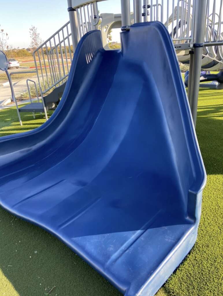 A large blue slide on a playground, set on artificial green turf. Other playground structures are visible in the background under a clear blue sky.