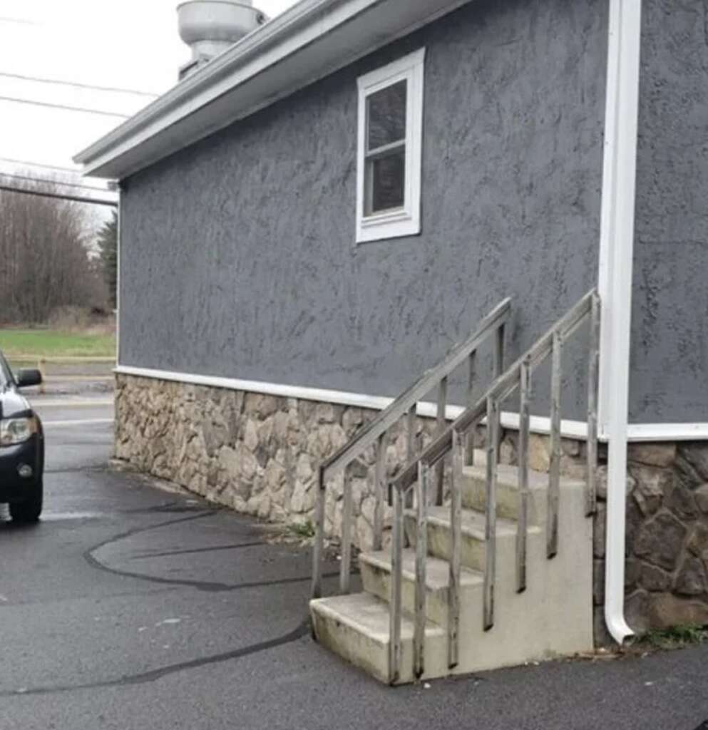 A small building with a gray wall, stone foundation, and a single window. Concrete steps with a metal railing lead to a door not visible in the image. A parked car is partially visible on the left side.