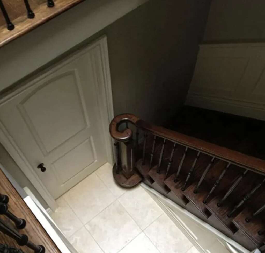 A staircase leading down between two walls, with a wooden handrail and dark-colored balusters. A closed white door with an arch near the bottom of the stairs. The floor is tiled with large, light-colored tiles.