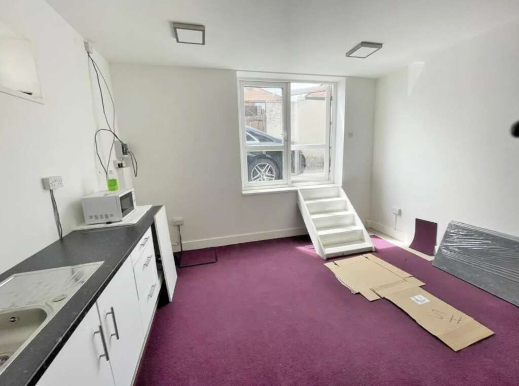 Small basement room with a purple carpet and a window showing part of a car outside. A white kitchenette with a sink, microwave, and some cables on the counter is on the left. Cardboard on the floor near the window, which has white steps leading up.