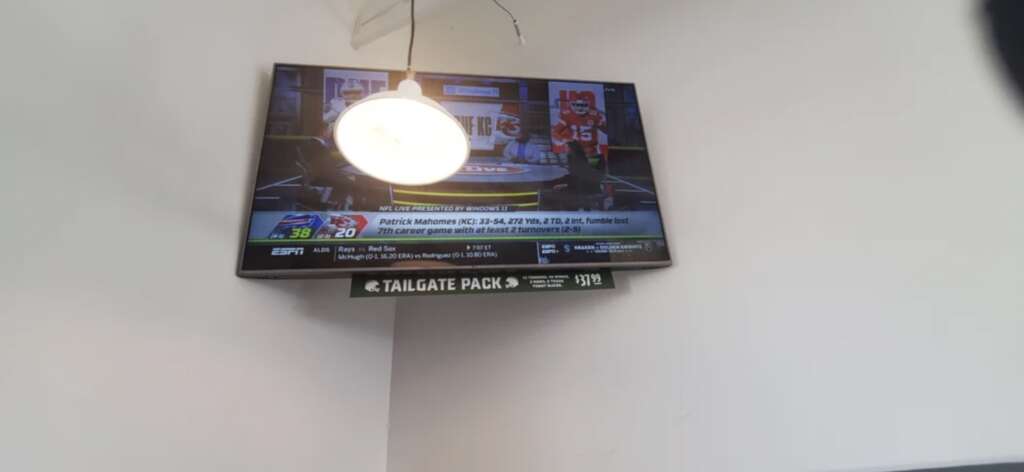 A wall-mounted TV displays a sports news channel, showing a football game update between two teams with scores 38 and 20. The screen is partially obscured by a ceiling light reflection. A sign below the TV reads "TAILGATE PACK.