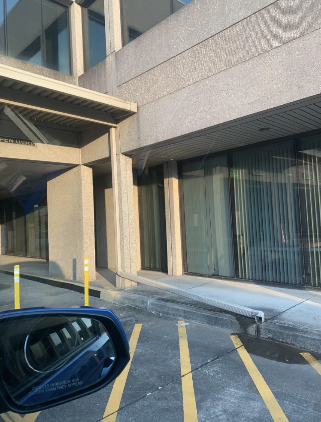 A parking lot with yellow striped lines leads to the entrance of a modern building with large windows and vertical blinds. A car's side mirror is visible in the foreground. The building exterior is made of textured concrete.