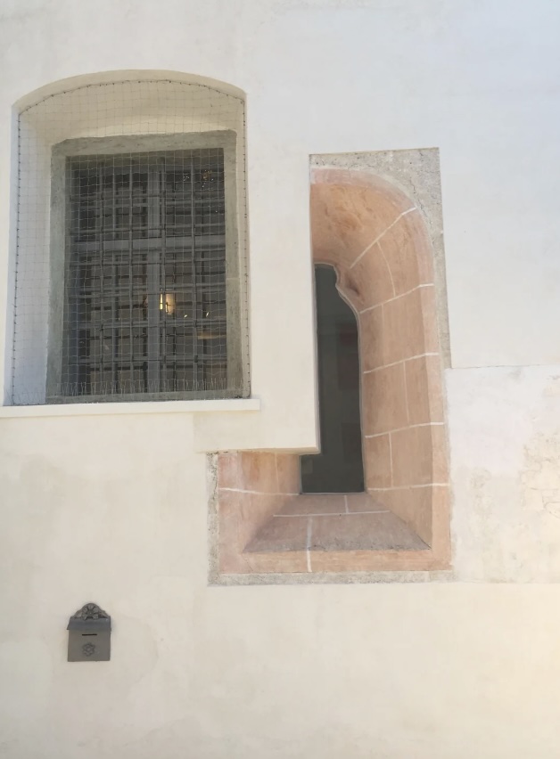 A pale, textured wall features two windows: one large with metal bars and netting, and a smaller one next to it with a stone frame. A small mailbox is mounted on the wall below the windows.