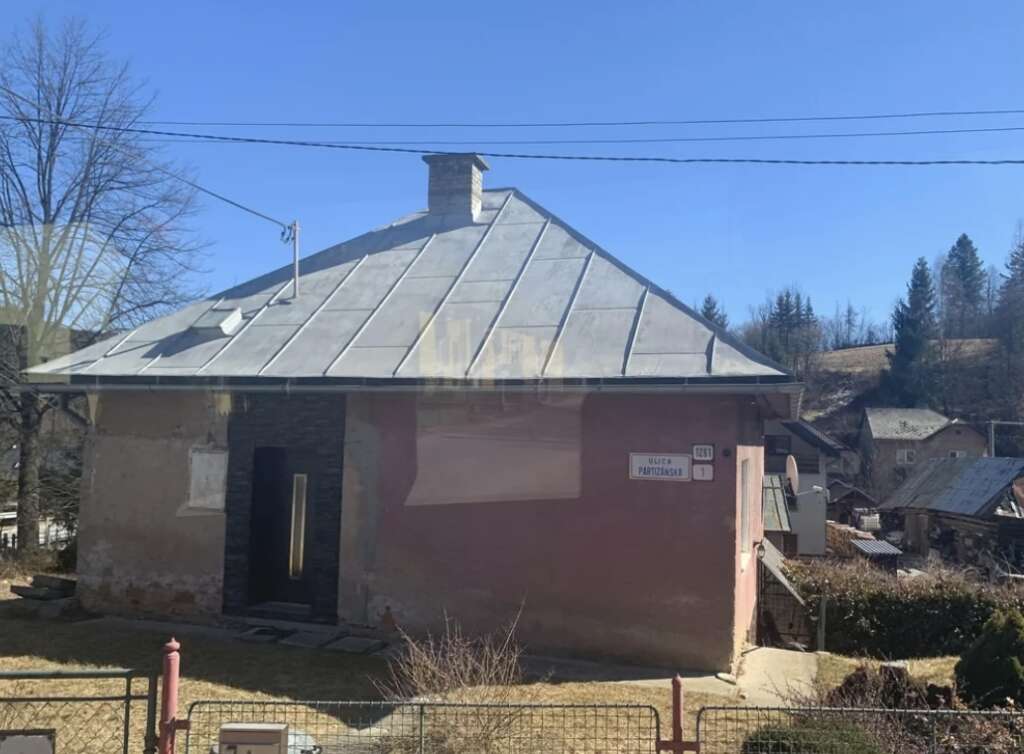 A small house with a metal roof stands on a grassy yard. It has a pinkish wall and a black door. There's a street sign on the corner of the building. Trees and more houses are in the background under a clear blue sky.