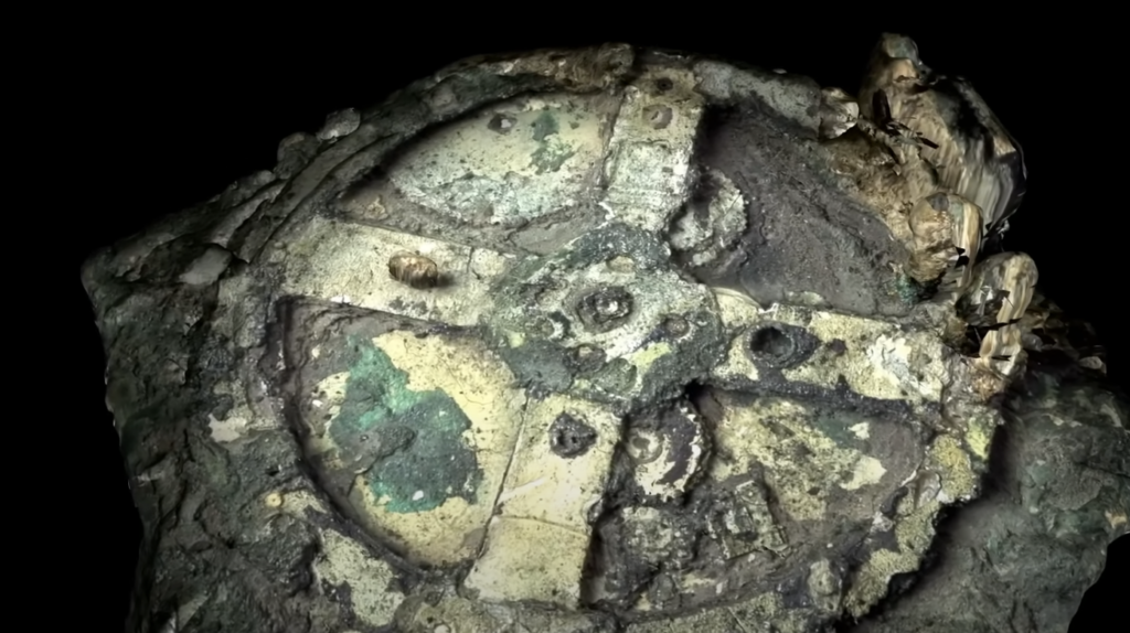 Close-up of the Antikythera mechanism, a corroded ancient Greek device, partially covered in green and brown patina. Its intricate gears and dials are visible, showcasing its complex design against a dark background.