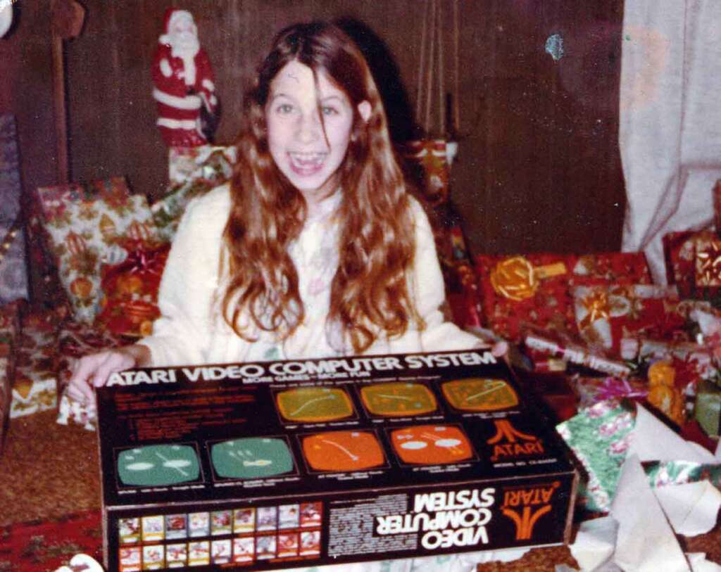 A girl with long hair excitedly holds an Atari Video Computer System box. She is sitting in a room with brown walls and Christmas decorations, including wrapped gifts and a Santa Claus decoration in the background.