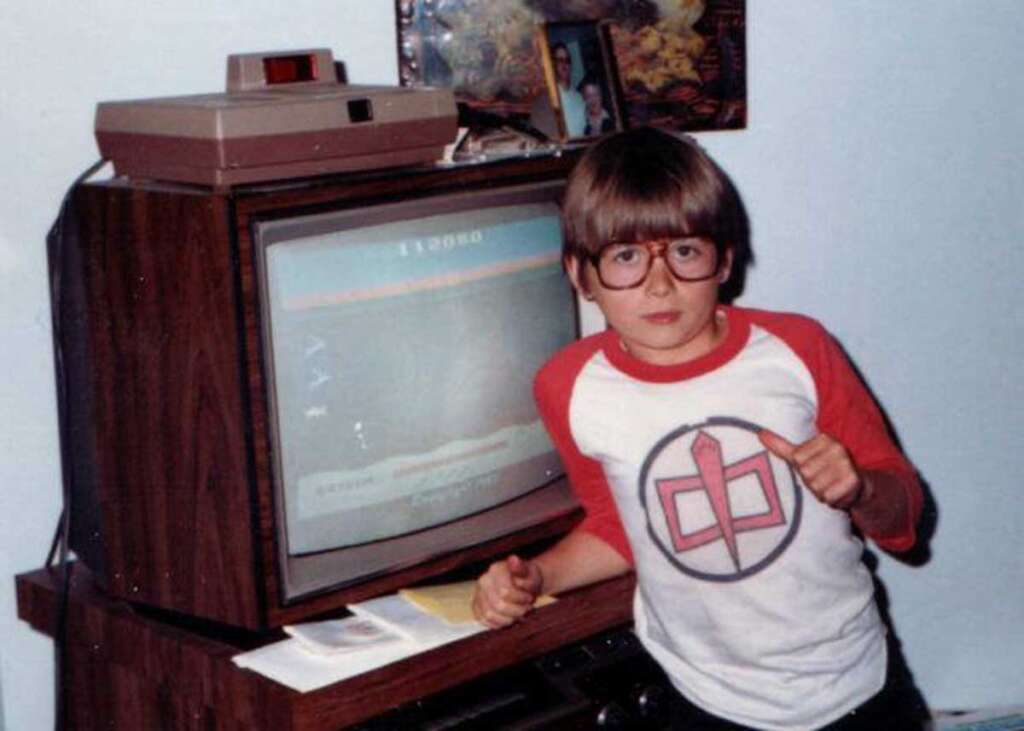 Young child with large glasses and a colorful t-shirt stands next to an old TV displaying a vintage video game. A game console is on top of the wooden TV cabinet, and various items are scattered around.