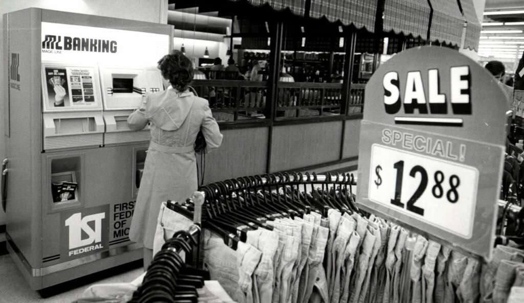 A person uses an ATM at "First Federal" near a clothing rack in a store. A "SALE" sign in the foreground advertises an item for $12.88. The background features more clothing and a counter with employees. The image is in black and white.