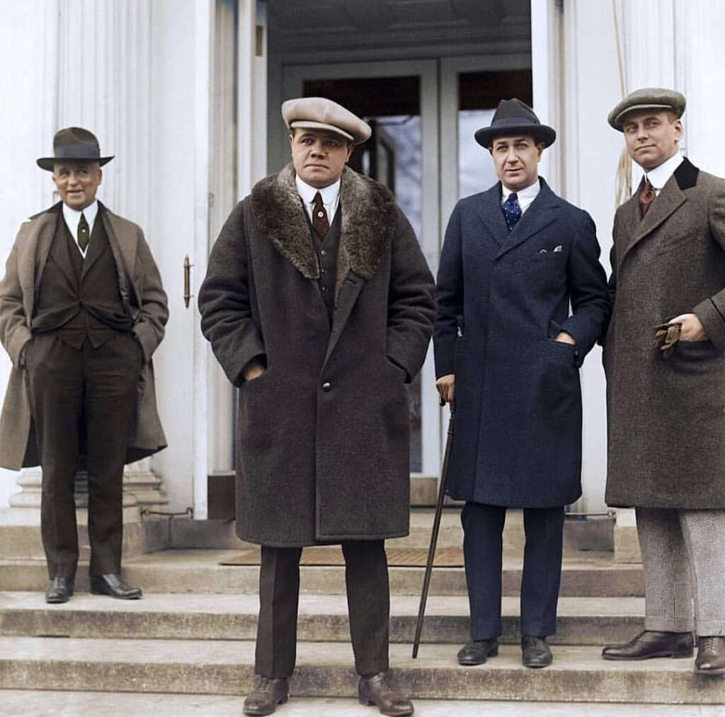 Four men in early 20th-century attire stand on steps in front of a building. They wear long coats and hats. One man holds a cane. The group appears formal and well-dressed.