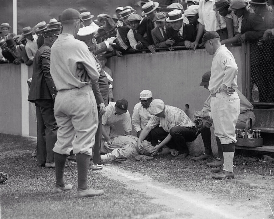 A group of baseball players and onlookers surround an injured player lying on the ground beside a fence. People in hats lean over the fence, watching the scene. Several men crouch and attend to the injured player.