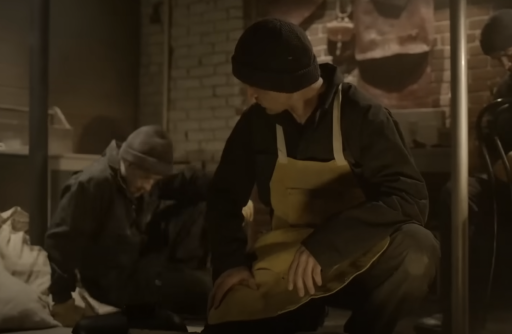 Two people wearing dark clothing and beanies work in a dimly lit room with brick walls. One person is crouching, wearing a yellow apron, while the other is seated nearby. Bags and tools are visible around them, suggesting a workshop setting.