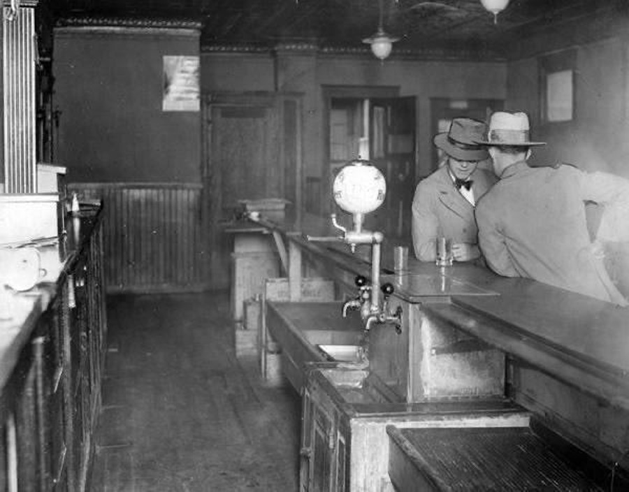 Two men in hats lean over a dark wooden bar in a dimly lit room, appearing to have a conversation. The bar is equipped with taps and there are bowls to the left. The room has a vintage, early 20th-century ambiance.