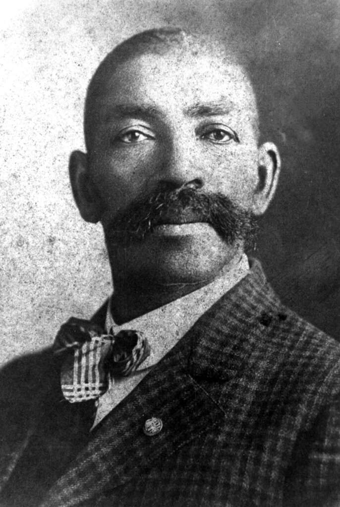 Black and white portrait of a man with a prominent mustache wearing a checkered suit, striped bow tie, and a pin on his lapel. The man is looking directly at the camera with a neutral expression.