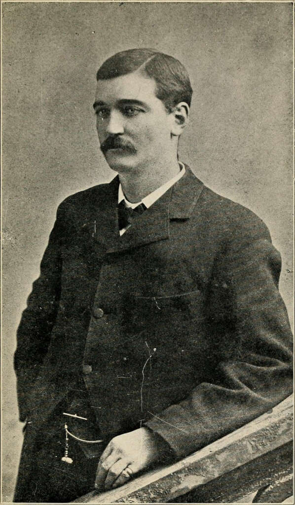 Black and white portrait of a man with short hair and a mustache, wearing a dark suit with a watch chain, leaning on a wooden rail with one arm. The man gazes slightly to the side. The background is plain.