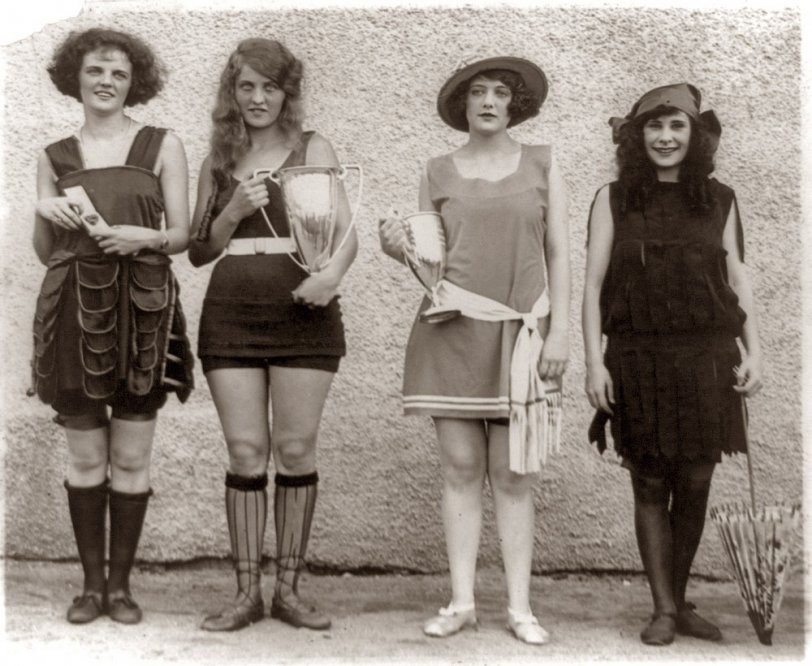 Four women in vintage swimsuits pose with trophies. They stand against a textured wall, each showcasing distinct fashion styles from the early 20th century, with one woman holding a parasol.