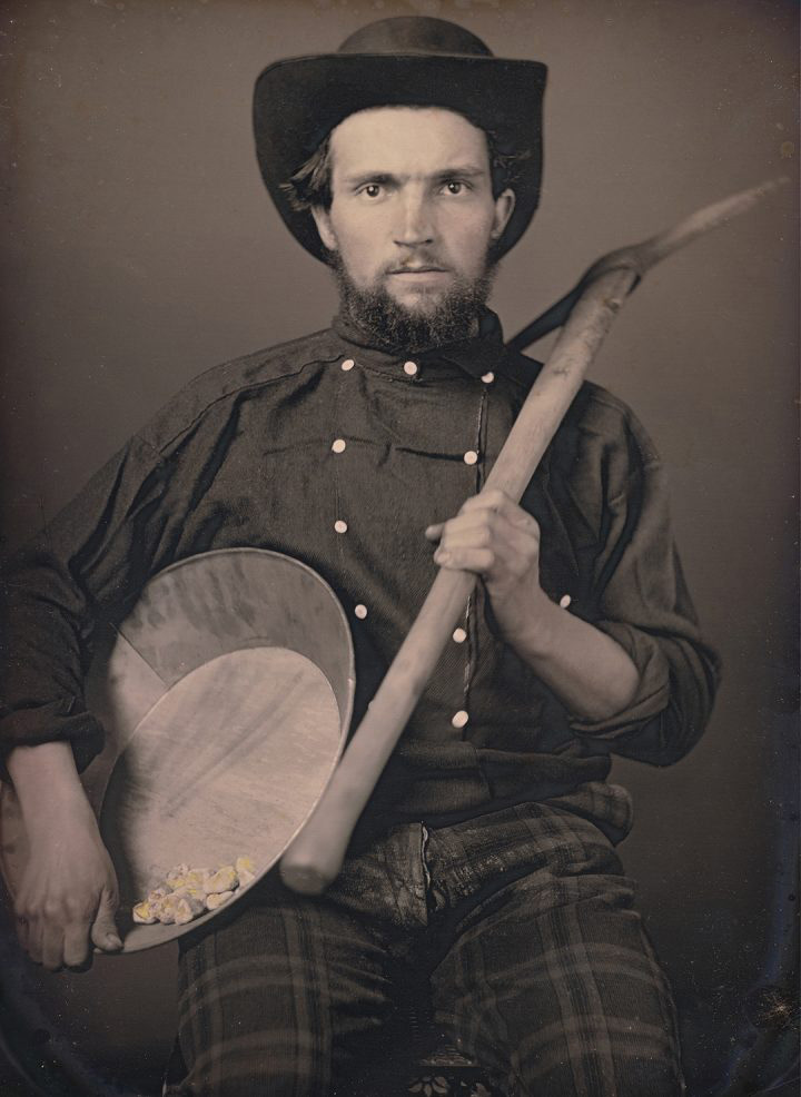 A man wearing a dark shirt and hat holds a pickaxe and a pan with gold nuggets. His expression is serious as he sits against a simple backdrop, evoking a historical or gold rush era setting.