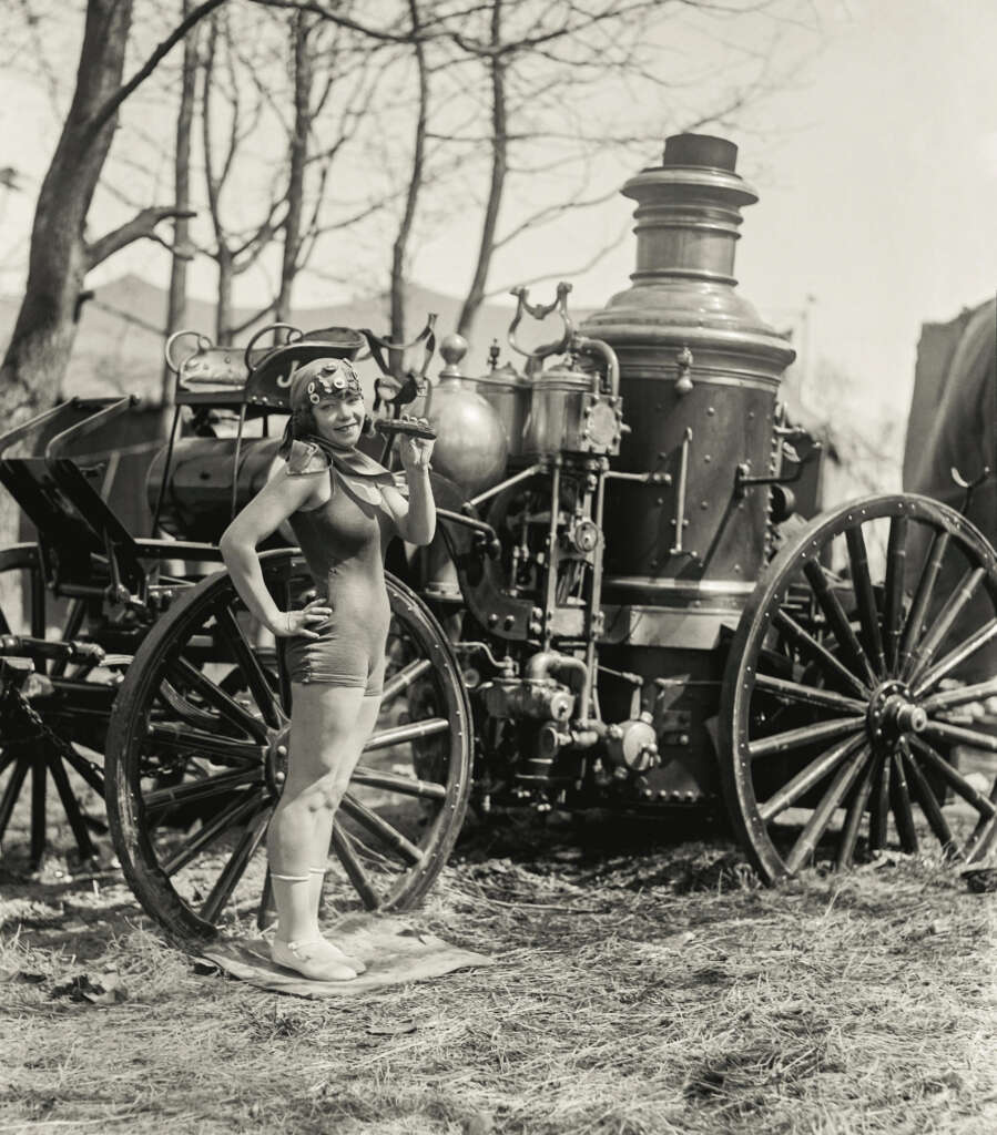 A woman in a vintage bathing suit poses with one hand on her hip and the other holding an ax, standing in front of an old steam-powered fire engine. Bare trees are visible in the background.