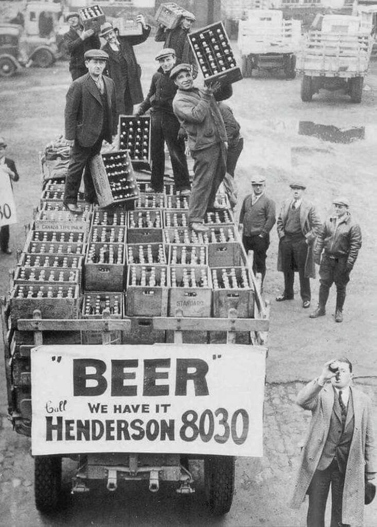 A vintage black-and-white photo showing men standing on a truck loaded with crates of beer bottles. The truck has a sign reading, "BEER - We have it," followed by a phone number. Other people stand nearby, with one man seemingly drinking.