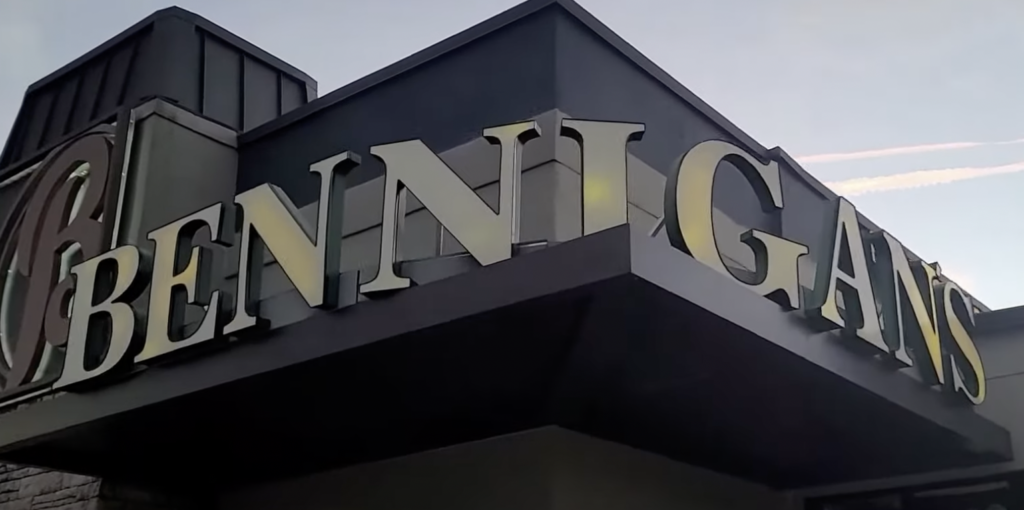 The image shows the sign for a "Bennigan's" restaurant. The letters are large and metallic, affixed to the corner of a building against a clear sky.