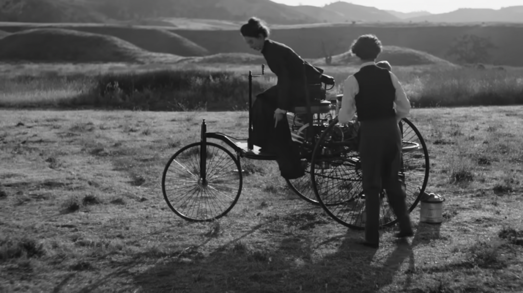 A black-and-white image showing two people with an early model three-wheeled motor vehicle in a field. One person is sitting on the vehicle while the other stands beside it, appearing to inspect or support it. Hills are visible in the background.