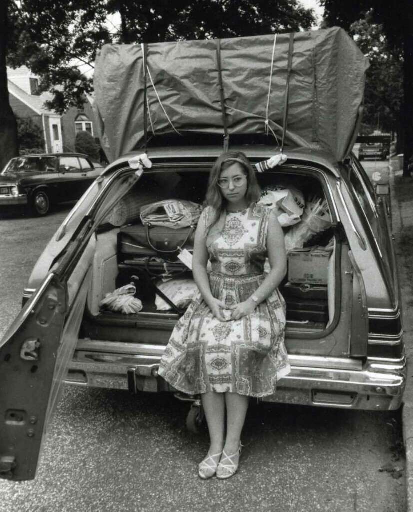 A woman in a patterned dress sits in the open trunk of a loaded station wagon. The car is packed with various items, and a large tarp is secured on the roof. Trees and vehicles line the street in the background.