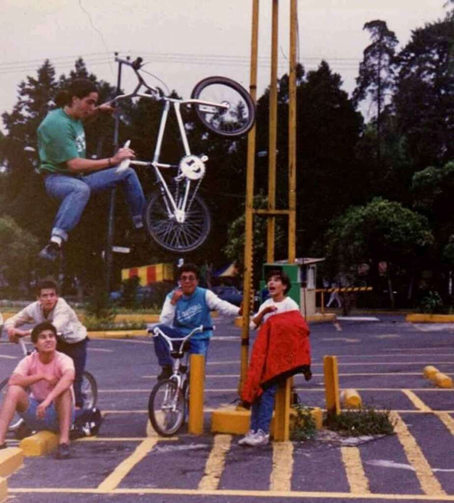 A person is performing a BMX trick in mid-air on a white bike, balancing on a yellow pole. Four other people look on from the ground, sitting on bikes or standing. The background features trees and a parking lot with yellow markings.