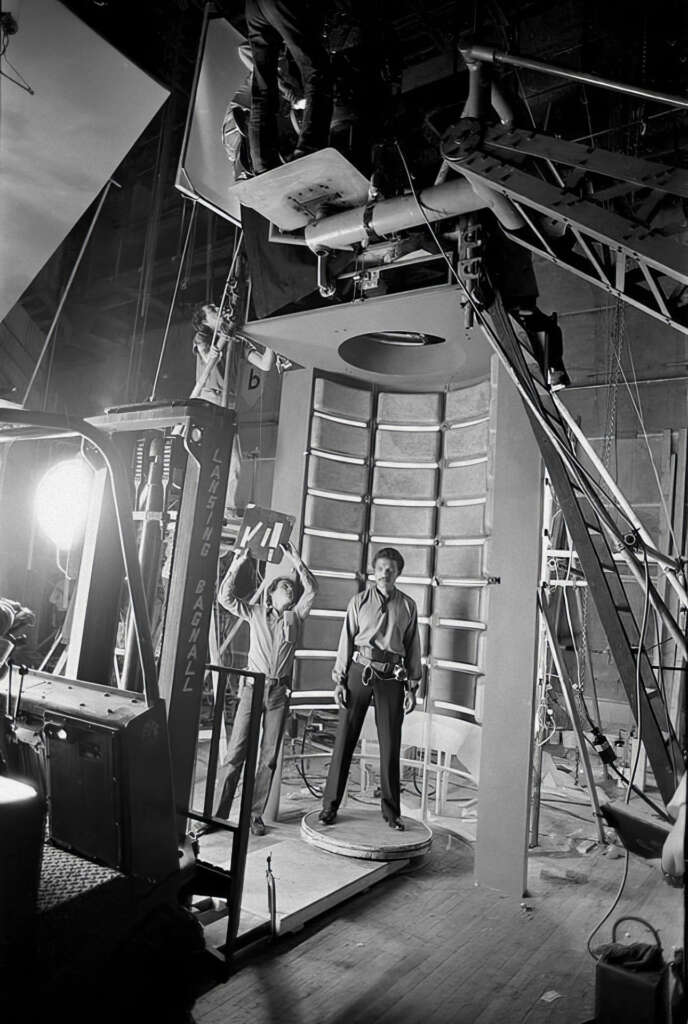 A person stands on a circular platform, surrounded by a large mechanical set with ladders and equipment. Crew members are positioned around, with lights and filming gear visible. The setting appears to be a film or TV production in a studio.