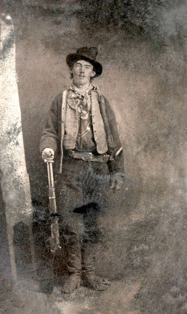 A sepia-toned vintage photo shows a person standing in cowboy attire, holding a rifle. They are wearing a hat, a jacket over a vest, and boots. The background is plain, giving an old-time Western feel.