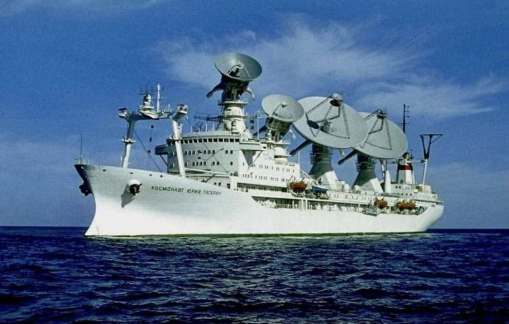 A white research ship with multiple large satellite dishes on its deck sails on a calm ocean under a clear blue sky.