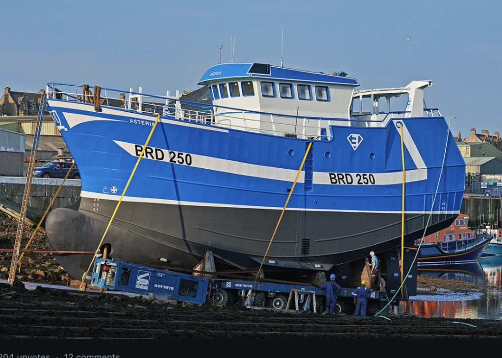 A blue and white fishing boat labeled "BRD 250" is elevated on a trailer with workers nearby. The scene is set in a harbor with buildings and another boat visible in the background.