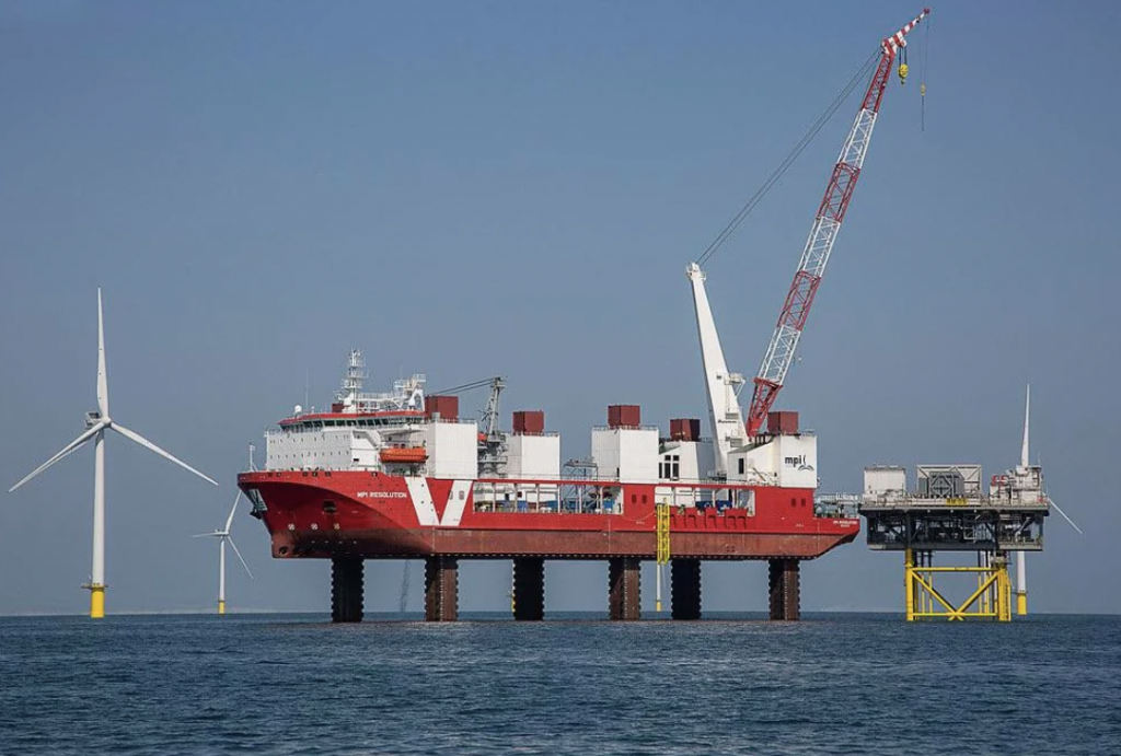 A large red and white offshore support vessel is stationed on elevated legs above the sea. It has a crane and is positioned near a wind turbine and a platform structure, under a clear blue sky.