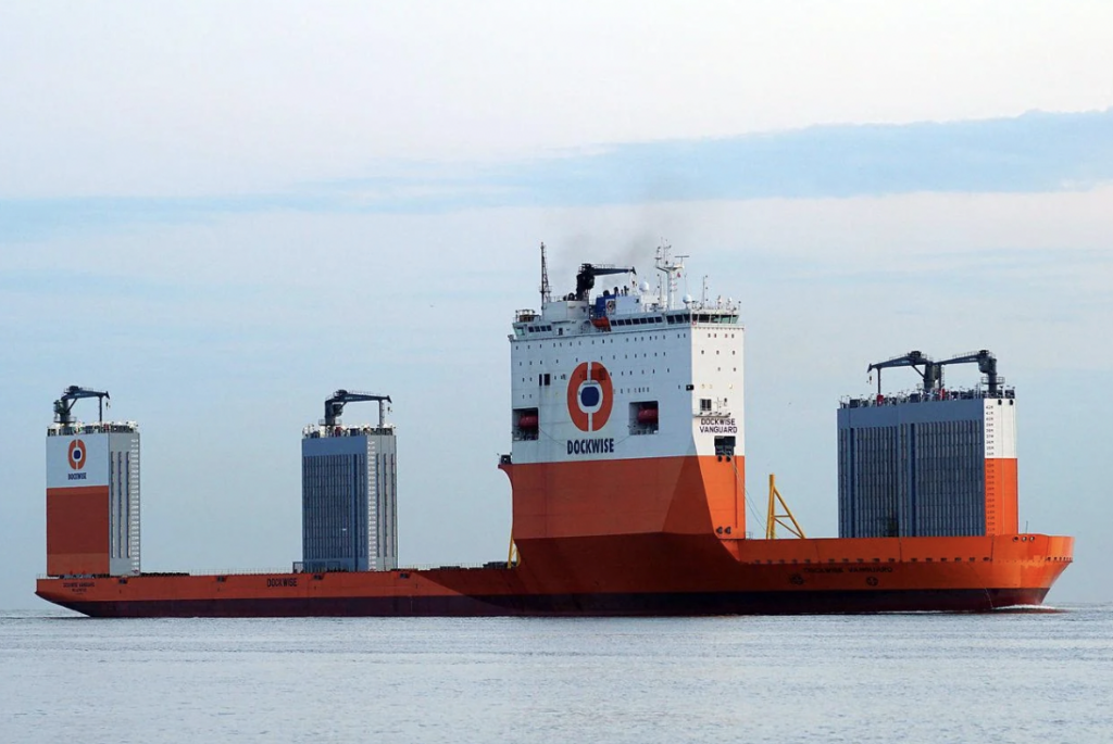 A large, specialized orange and white heavy-lift vessel with the name "Dockwise" on its side sails on calm waters. The ship has a flat deck and tall structures resembling gantry cranes on both sides. The sky is clear with a few clouds.