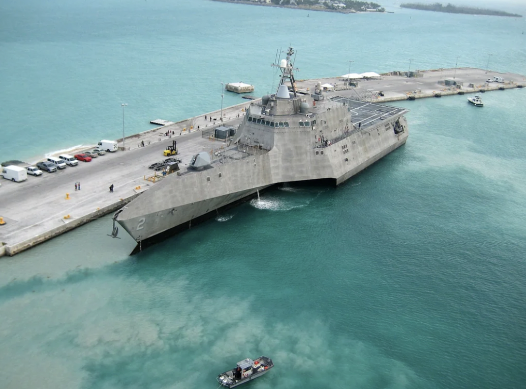 A modern naval ship docked at a pier with a sleek, gray design and angular features. The surrounding water is turquoise, and a small boat is nearby. Several vehicles and people are visible on the pier in the background.